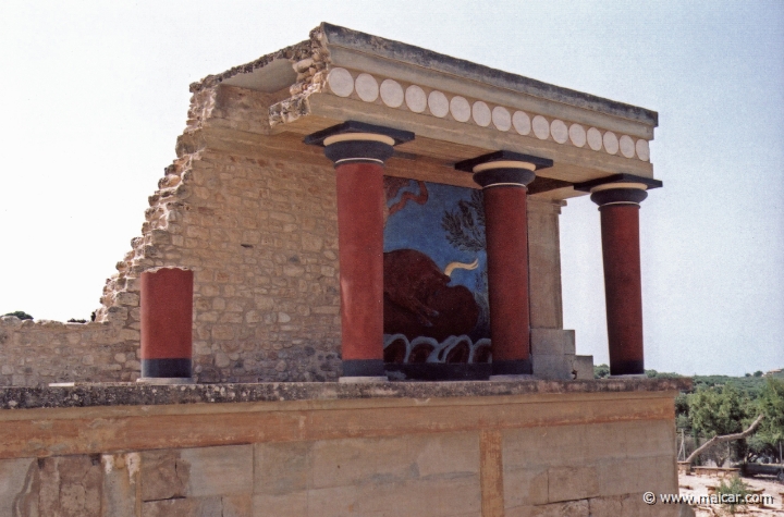 9532.jpg - 9532: Bull Bastion on the west side of the Palace of Knossos. An open air passage linked the Central Court with the North Entrance. It was paved and sharply inclined towards the north. The passage is narrow. Right and left were two raised colonnades known as “Bastions.” Arthur Evans reconstructed the “Bastion” on the west side. He also placed a copy of a restored relief fresco of a bull here. The wall painting may have formed part of a hunting scene. The passage ends in a large hall with ten square pillars and two columns. The pillars and columns probably supported a large hall on the upper floor. Evans suggested that, due to its position on the seaward side, it was here that the produce of seaborne trade would have been checked when it reached the Palace. It was therefore named the “Customs House”. Palace of Knossos (Crete).