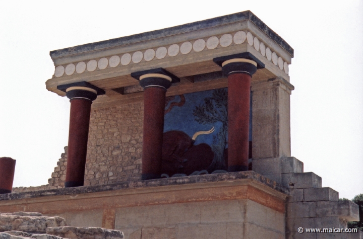 9535.jpg - 9535: Bull Bastion on the west side of the Palace of Knossos. An open air passage linked the Central Court with the North Entrance. It was paved and sharply inclined towards the north. The passage is narrow. Right and left were two raised colonnades known as “Bastions.” Arthur Evans reconstructed the “Bastion” on the west side. He also placed a copy of a restored relief fresco of a bull here. The wall painting may have formed part of a hunting scene. The passage ends in a large hall with ten square pillars and two columns. The pillars and columns probably supported a large hall on the upper floor. Evans suggested that, due to its position on the seaward side, it was here that the produce of seaborne trade would have been checked when it reached the Palace. It was therefore named the “Customs House”. Palace of Knossos (Crete).